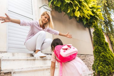 Kolları açık olan mutlu gülümseyen anne öğrenci kızı evde anaokulundan dönecek karşılamaktadır. İyi bir ilişki. Mutlu sevimli küçük kız annesi ile okul öncesi günün ardından mutlu hissediyorum.