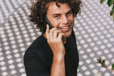 Genel gider closeup mutlu yakışıklı genç adam açık havada cep telefonda konuşurken ayakta siyah t-shirt giyen portresi. Arkadaşıma telefon görüşmesi gülümseyen ve kamera için arayan genç adam