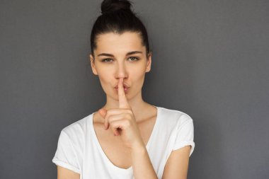 Esmer kendine güvenen erkek portresi beyaz T-shirt giyiyor, gösterir sessizlik işareti, bilgileri gizli tutmak sorar vardır ciddi ifade, gri studio arka plan üzerinde izole. Gizli jest, shhh.