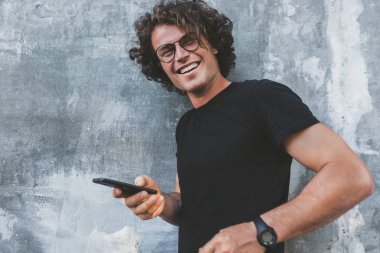 gri duvara karşı duran ve kameraya seyir sırasında cep telefonu tutan kıvırcık saçlı yakışıklı erkek gülümseyen