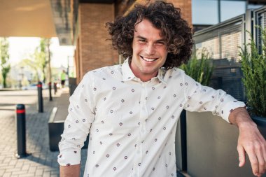 Genç neşeli erkek modeli kıvırcık saçlı ve çilli, gülümseyen portresi açık kafe poz ve dişlek gülümseyerek kameraya bakarak trendy gömlek giyer. Kişi, yaşam tarzı ve iş kavramı.