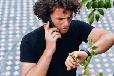 Ciddi genç adam açık havada hareket eden telefon için saati görünümlü konuşurken ayakta closeup görüntüsü. Yakışıklı deli erkek telefon görüşmesi ve onun kız arkadaşı bekliyor. İnsanlar ve yaşam tarzı kavramı