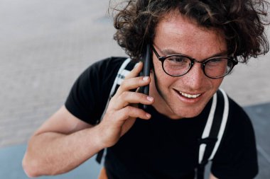 yukarıdan açık havada cep telefonunda mesaj atıp messsages oturan yakışıklı mutlu adamın görüntüleyin. Kıvırcık saçlı Genç erkek ücretsiz kablosuz akıllı telefon kullanarak şehirde dinlenme gözlük takıyor. 