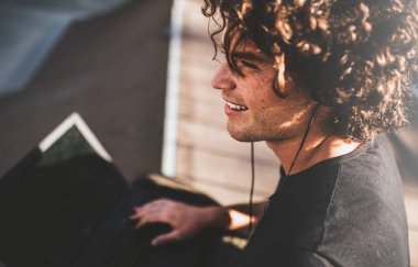 Closeup arka sohbet için online ile sokulgan ve müzik için ücretsiz kablosuz bağlı kulaklık dizüstü kullanarak kıvırcık saçlı gülümseyen erkek görüntüsünü görüntüleyin. Yakışıklı adam manifatura iletileri.