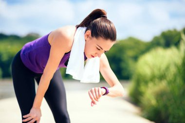 Çekici kadın önce veya sonra egzersiz ve parkta çalışan açık yapma. Mor t-shirt ve beyaz havlu ile doğa arka plan üzerinde. Onun çalışma süresi kontrol saate bak. Spor