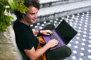 Şehrin sokak üzerinde ücretsiz kablosuz dizüstü online arkadaşlarınızla sohbet için kullanma kıvırcık saçlı erkek kadeh bağlı görünümü arka. Yakışıklı hipster adam manifatura kendi dizüstü bilgisayarında bir şey.