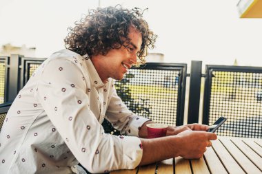 Ücretsiz wifi akıllı telefon üzerinden tarama şık gömlek, kıvırcık saçlı yakışıklı erkek gülümseyen Closeup yan görünümü portresi. Çekici bir adam manifatura kafe Açık iletide. İnsanlar, yaşam tarzı, teknoloji
