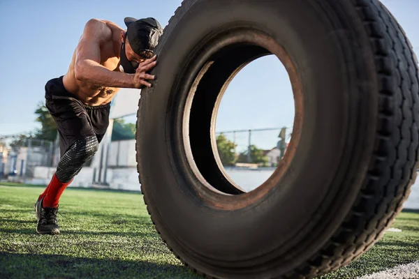 Attractive fitness muscular man in mask to increase load on breathing muscles doing exercises with tire outdoors. Shirtless sportsman doing workout on stadium green grass. Sport and people