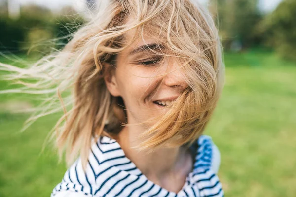 Cropped close up outdoor shot of attractive happy female with blond blowing hair and closed eyes enjoy weather. Close up shot of beautiful young woman smiling with windy hair in park. People concept