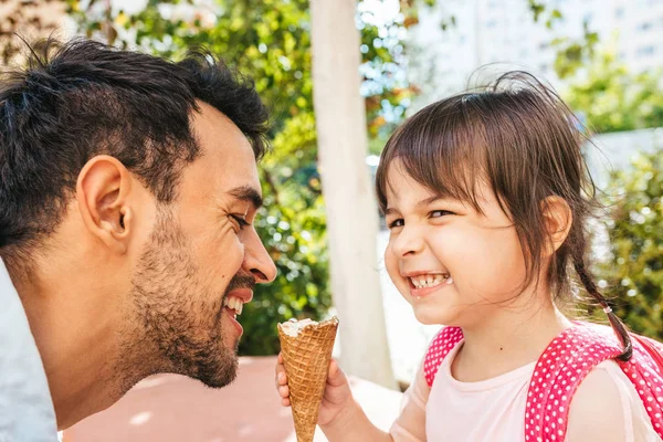 Closeup side view of happy cute little girl sitting with handsome dad eating ice-cream outdoors. Fun girl kid and cheerful father have fun and enjoy outside. Good relationship between dad and daughter