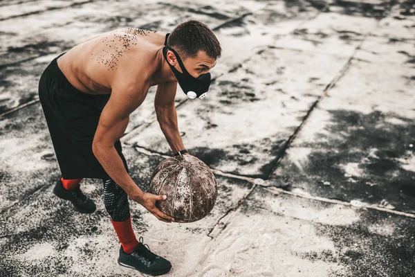 Athlete muscular male doing exercises with medicine ball in mask on concrete background outdoor with copy space for your advertising. Caucasian shirtless sportsman doing workout