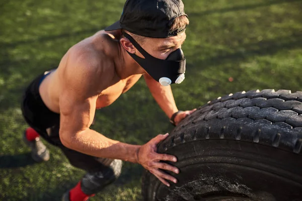 Closeup portrait of Caucasian fitness muscular man in mask to increase load on breathing muscles doing exercises with tire outdoors. Shirtless sportsman doing workout on stadium green grass. Sport