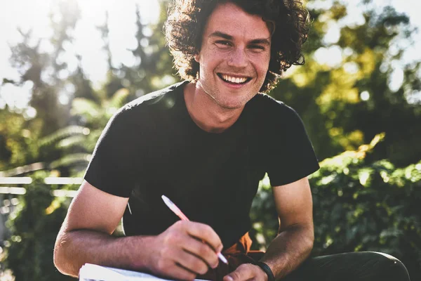 Joyeux Jeune Homme Souriant Aux Cheveux Bouclés Écrivant Quelques Notes — Photo