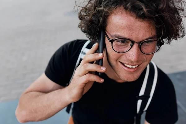 view from above of handsome happy man sitting outdoors texting messsages on mobile phone. Young male with curly hair wears spectacles resting in the city using free wireless on smart phone.