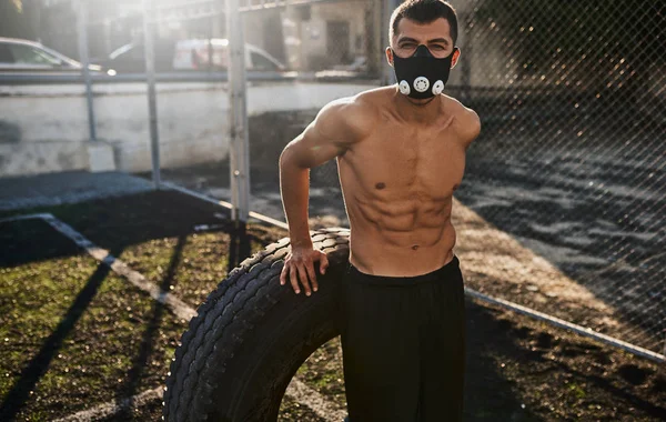 Horizontal portrait of muscular male posing in mask to increase load on breathing muscles doing exercise with big tire outdoors in stadium. Copy space for text. Shirtless sportsman doing workout