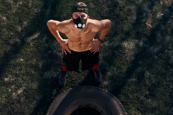 View from above of muscular young male with mask looking at the camera while doing exercises with big tire outdoors in stadium. Shirtless sportsman doing hard workout, healthy lifestyle. People, sport