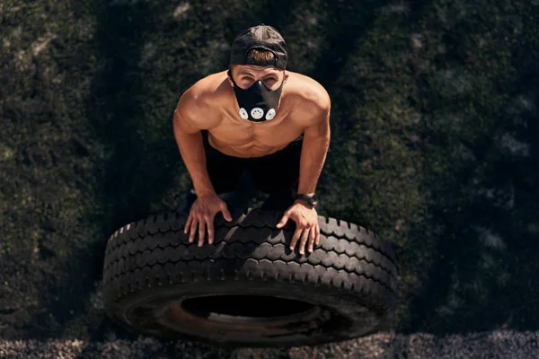View from above of muscular young male with mask doing exercises with big tire outdoors in stadium. Shirtless sportsman doing hard workout, healthy lifestyle. People and sport concept.