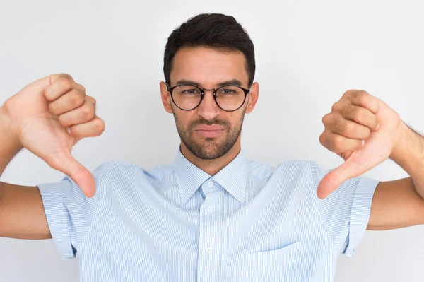 Portrait of unhappy male wears round trendy glasses, striped shirt looking at the camera with negative expression, showing thumbs down with both hands. Body language and emotion concept