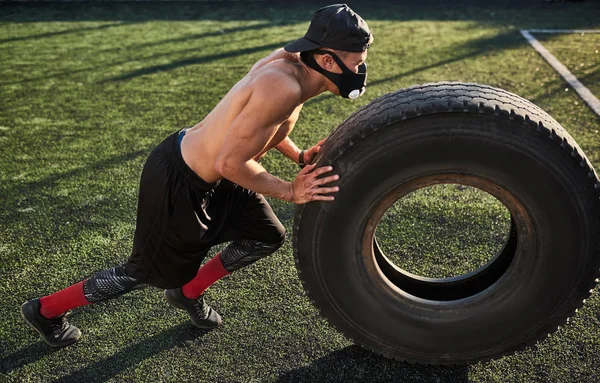 Above view shot of Caucasian fitness muscular man in mask to increase load on breathing muscles doing exercises with tire outdoors. Shirtless sportsman doing workout on stadium green grass. Sport