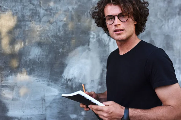student man with curly hair posing on camera at grey wall and holding book