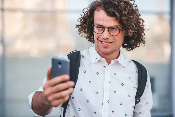 漂亮的年轻迷人的男性穿着时髦的眼镜和休闲衬衫 玩得很开心 在户外用智能手机自画像 走在大街上 生活方式和技术理念 — 图库照片