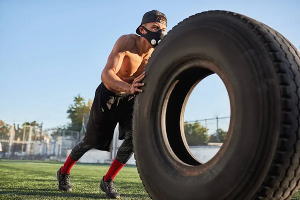 Caucasian fitness muscular man in mask to increase load on breathing muscles doing exercises with tire outdoors. Shirtless sportsman doing workout on stadium green grass. Sport and people