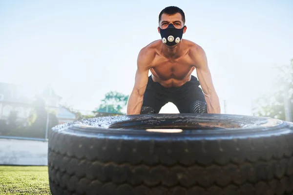 Outdoor shot of young athlete male in mask to increase load on breathing muscles doing exercise with big tire outdoors in stadium on sunlight. Copy space. Shirtless sportsman doing workout, healthy