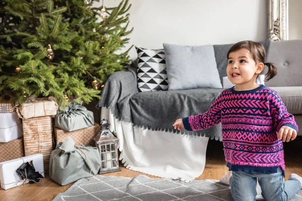 Happy surprised toddler girl to see a lot holiday gifts under Christmas tree. Merry Christmas and Happy New Year! Cute little child girl choose the present. Beautiful girl and Christmas tree indoors.