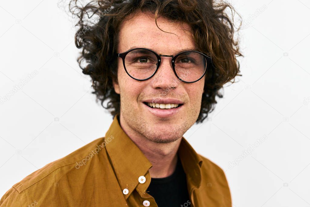 handsome man with curly hair, wearing glasses and posing and standing against white wall