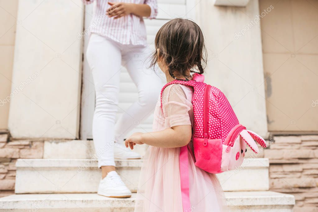 Image of mother standing on the stairs against the home saying goodbye to her daughter as she leave for kindergarten. Cute little girl wears dress and backpack going to preschool, waving to mother 