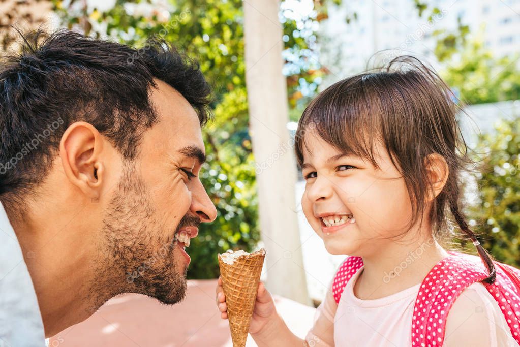 Closeup side view of happy cute little girl sitting with handsome dad eating ice-cream outdoors. Fun girl kid and cheerful father have fun and enjoy outside. Good relationship between dad and daughter