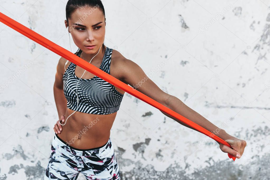 Horizontal portrait of fitness woman standing against concrete wall doing stretching exercises holding a red resistance band. Athletic female listen the music on earphones during workout outdoor.Sport