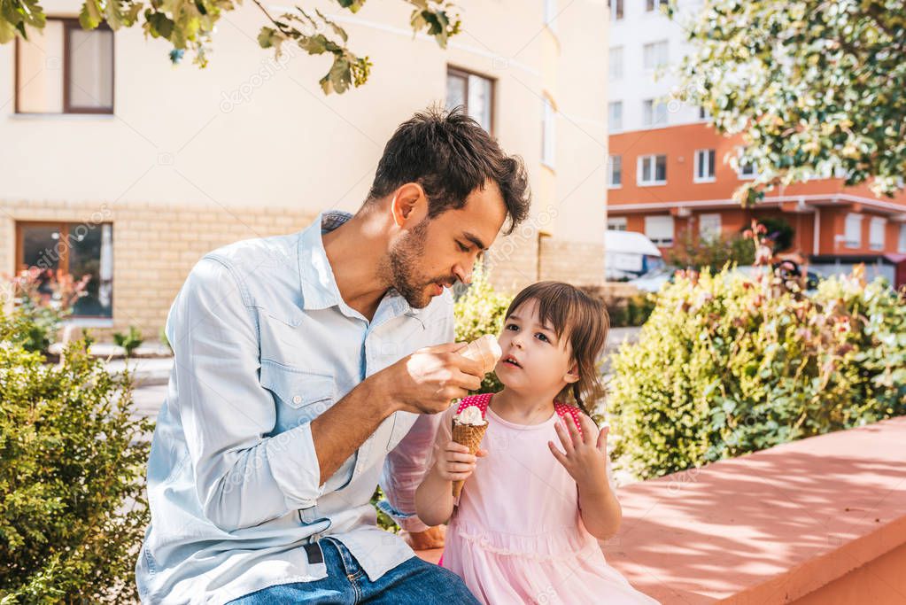 Horizontal portrait of little girl sitting with dad on the street and eating ice-cream outdoor. Happy kid wears dress spending time together with father. Good relationship between dad and daughter