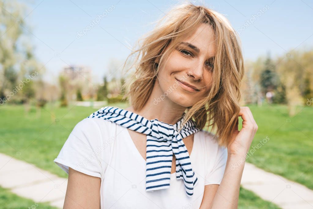 Outdoor portrait of beautiful female with blonde blowing hair, smiling and looking at the camera. Closeup view of Caucasian young woman smile with hand on windy hair in park. People, lifestyle concept