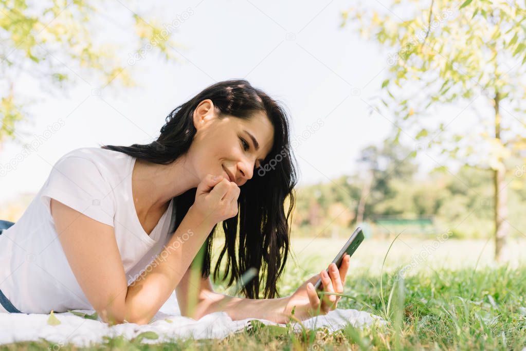 Horizontal portrait of smiling young beautiful female having happy look while talking on mobile phone, recieving good news, sitting at the park on white blanket and green grass. People and lifesyle.