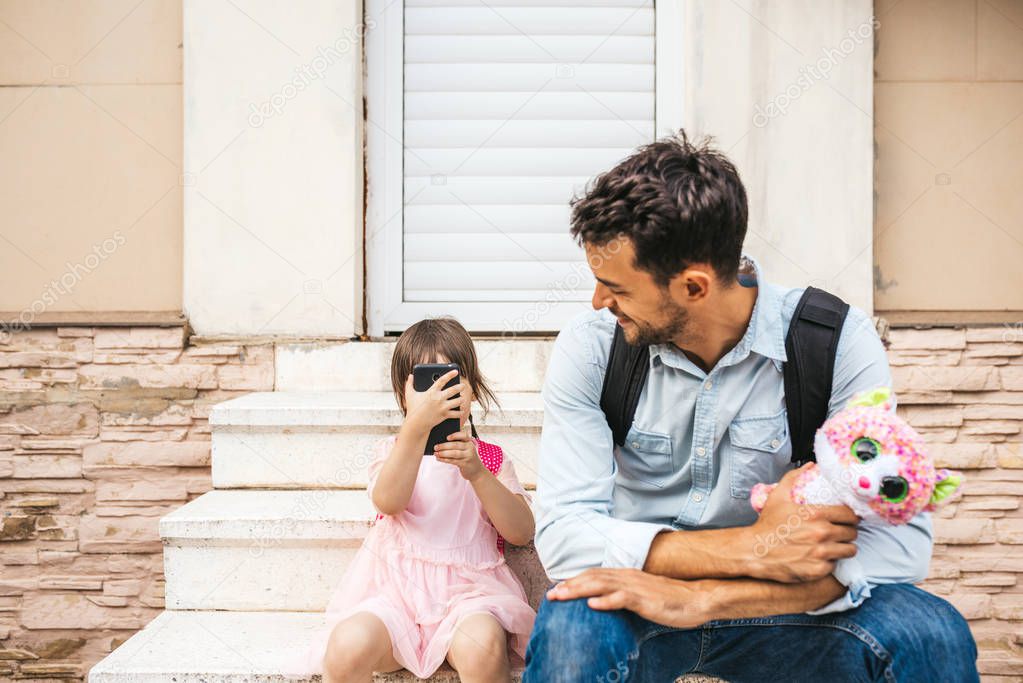 Handsome young father spending the time together with his daughter pupil after preschool. Cute little girl play smart phone taking photos with his dad. Both are sitting against of home on the stairs.