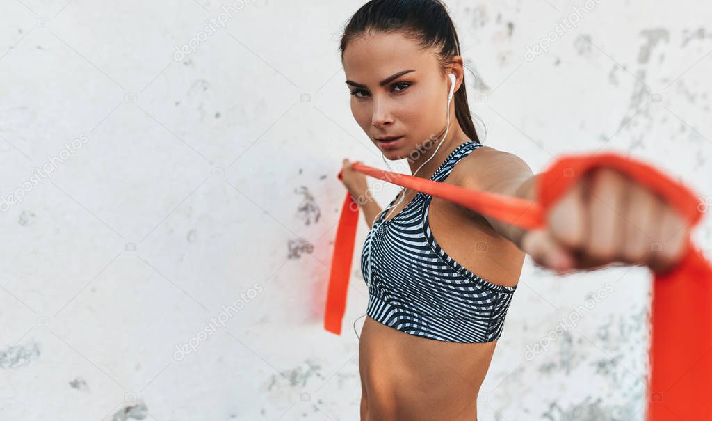Closeup portrait of fitness woman standing against concrete wall doing stretching exercises holding a red resistance band. Athletic female listen the music on earphones during workout outdoor.