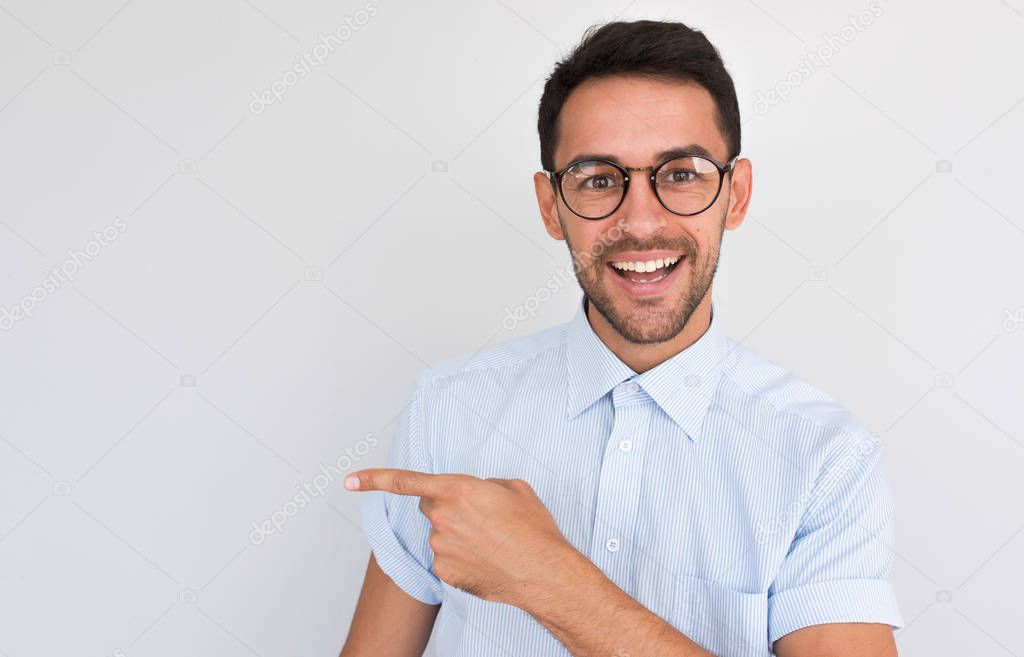 Portrait of happy unshaven handsome male smiling with toothy smile, points aside, looking to the camera, shows copy space for your advertising content, against white studio wall, in casual blue shirt.
