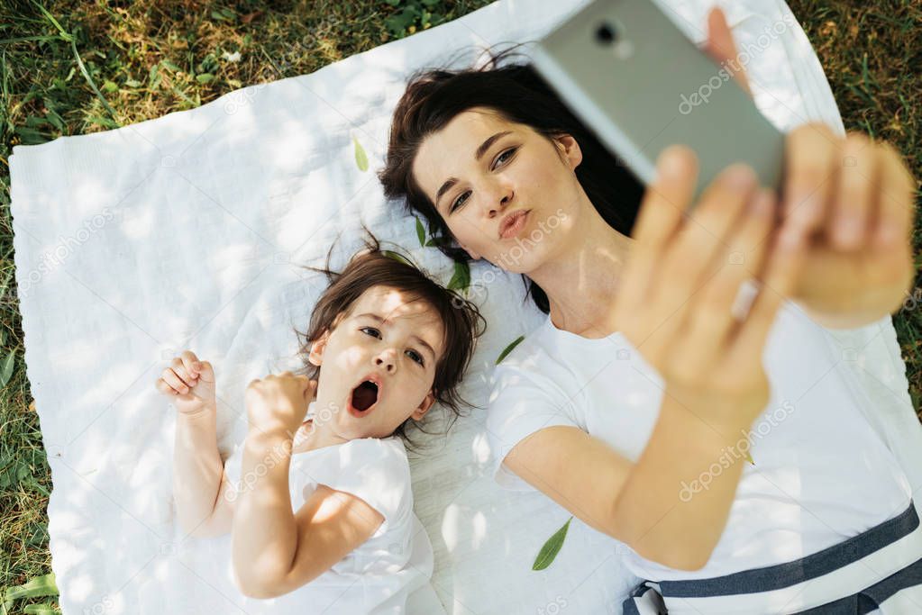Horizontal portrait of young beautiful woman mother blowing kiss and taking self portrait with her cute kid lying on green grass and white blanket. Motherhood, technology and childhood concept.