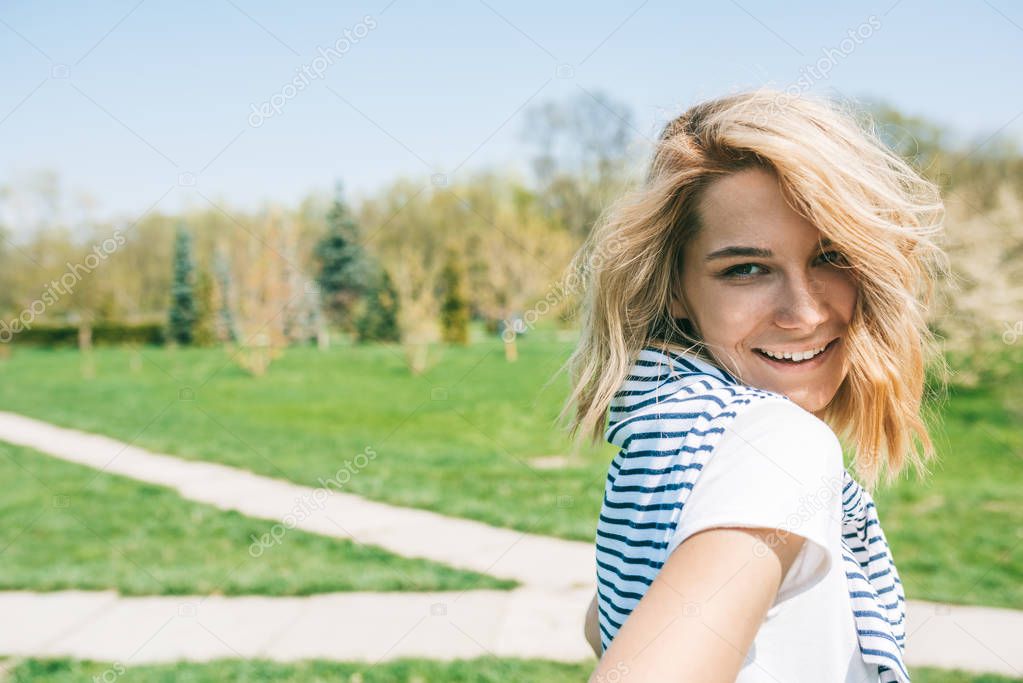 Closeup portrait of attractive happy female with blonde hair, smiling and posing against nature background. Portrait of Caucasian young beautiful woman smile with windy hair in park. People concept