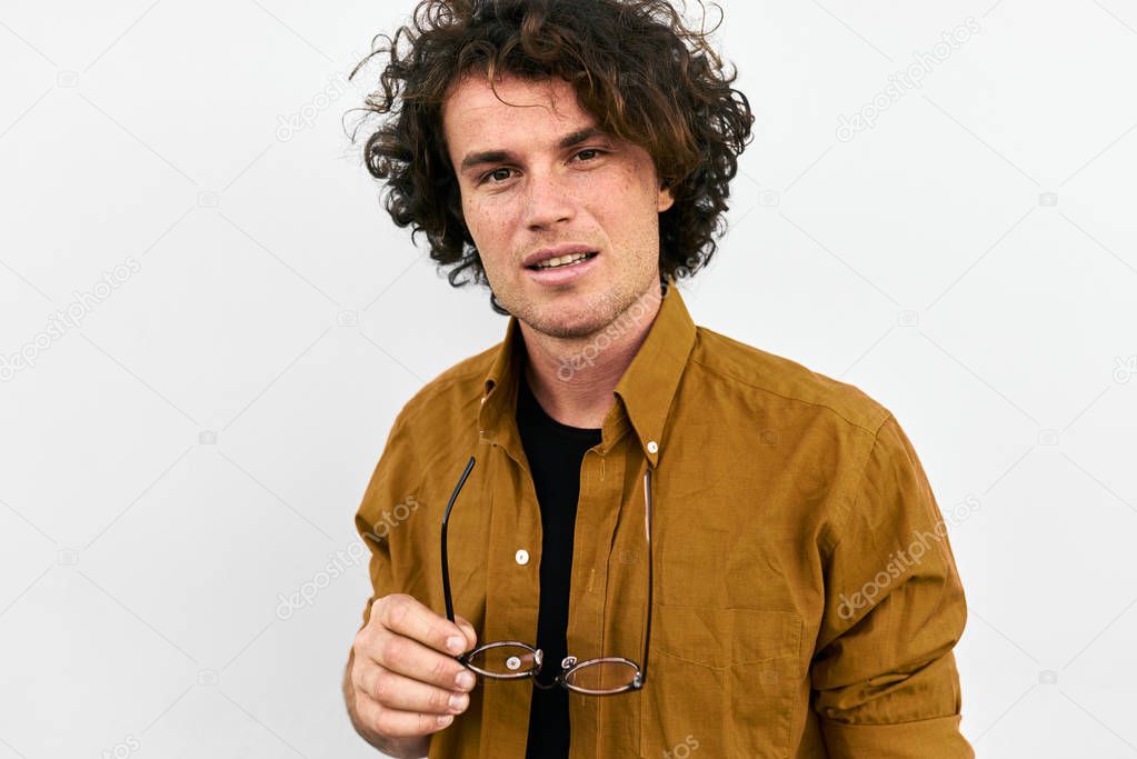 Horizontal closeup portrait of handsome smart man with curly hair, with spectacles in hand posing for social advertisement, isolated on white wall with copy space for your promotional information. 