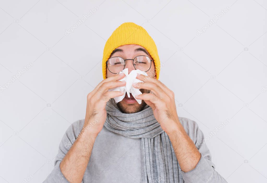 Closeup portrait of sick handsome man wearing grey sweater, yellow hat and spectacles, blowing nose into handkerchief. Male have flu, virus or allergy respiratory. Healthy, medicine and people concept