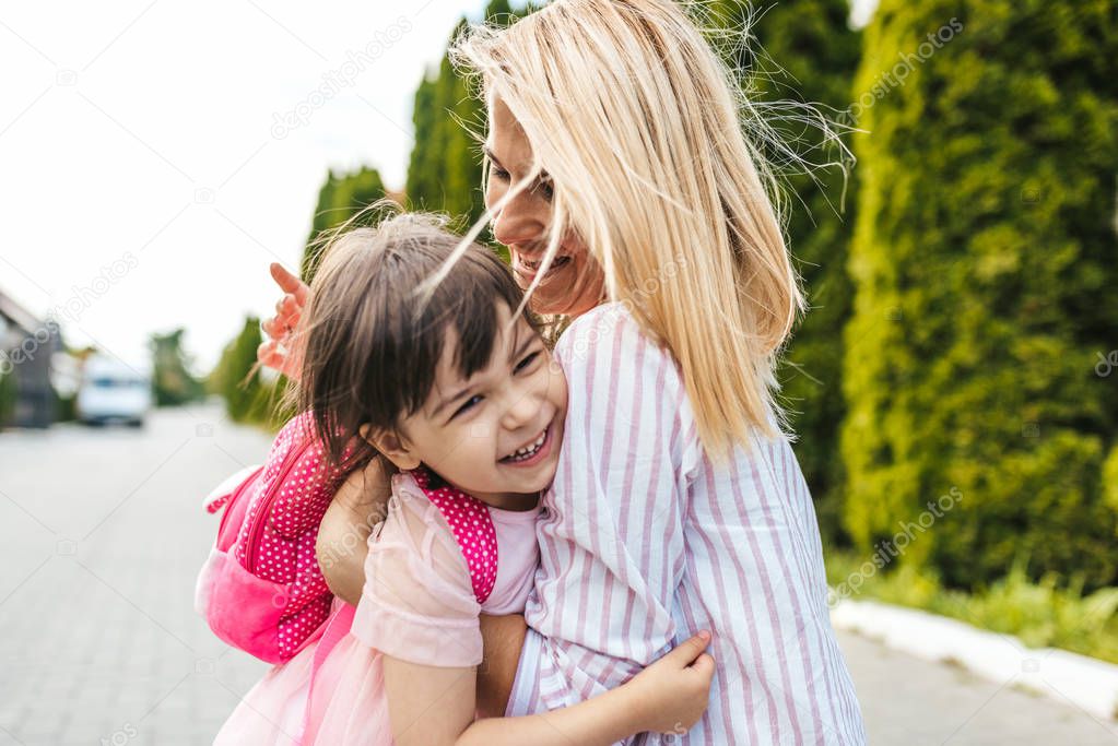 Happy little girl and pretty mother embracing each other on sidewalk next to the trees. Mom play together with kid preschooler with backpack outdoor. Mom and child have fun. Mother's day concept.