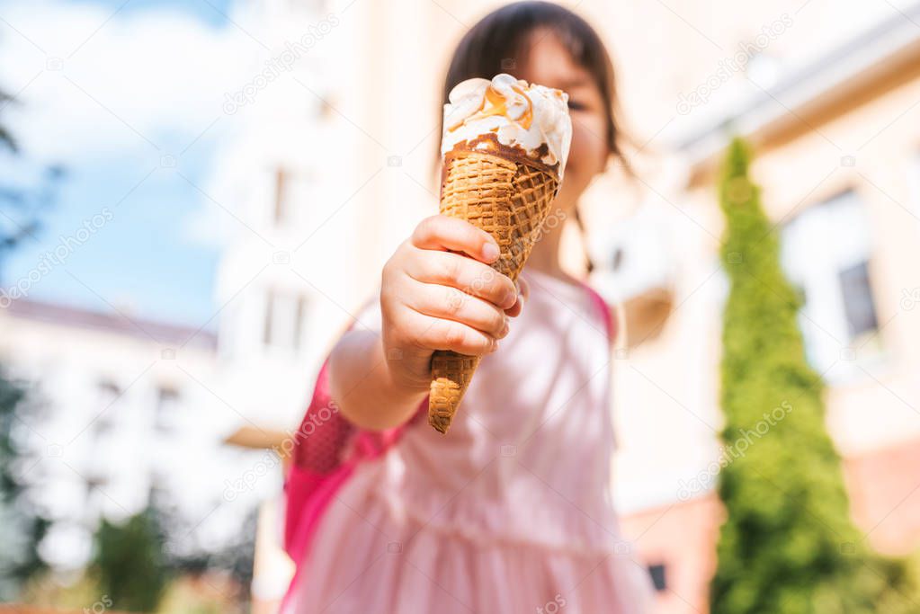 Closeup image of cute little girl walking along city street and eating ice cream outdoor. Happy kid girl showing her ice-cream in focus with blurred background. Happy preschooler with pink backpack.