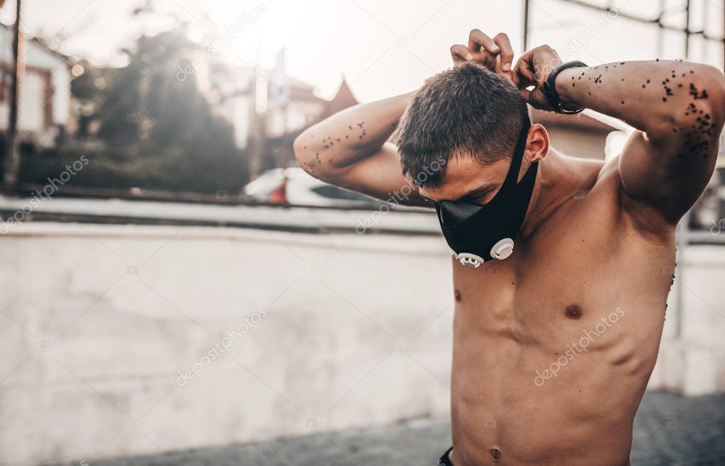 Caucasian muscular male wearing mask to increase load on breathing muscles during morning cardio workout routine outdoors. Copy space. Shirtless sportsman doing workout outside