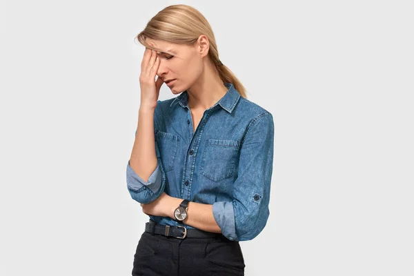 Tired young female touching her head with closed eyes posing on white studio background. Caucasian blonde business woman wearing denim shirt, suffering from migraine. People, business, health concept