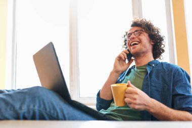 Bir dizüstü bilgisayarda çalışırken cep telefonu üzerinde konuşurken ev ofis içinde oturan genç serbest meslek sahibi adam. Dizüstü bilgisayarda çalışma, cep telefonunuzun konuşma ve bir kahve içmek işadamı gülümseyen.