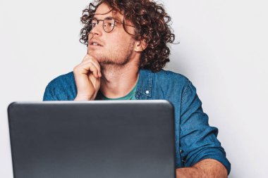 Kıvırcık saçlı laptop çalışma ve düşünme masada oturan genç ciddi adam. Yuvarlak gözlük takan ve dizüstü bilgisayar üzerinde çalışan iş adamı.