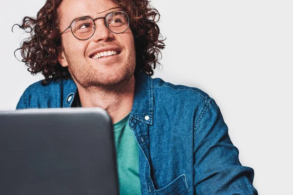 Крупним планом студійний знімок молодого усміхненого чоловіка з кучерявим волоссям, що сидить за столом, працює на ноутбуці та мріє. Бізнесмен носить круглі окуляри і працює на портативному комп'ютері . — стокове фото