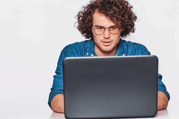 Obrázek z vážných kavkazských podnikatel pracuje na jeho laptopu s kudrnatými vlasy a nosí ležérní modrou košili. Pohledný mladý muž u stolu, Pózování na studio pozadí. — Stock fotografie